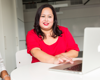 Woman in red top and lipstick sat by computer smiling